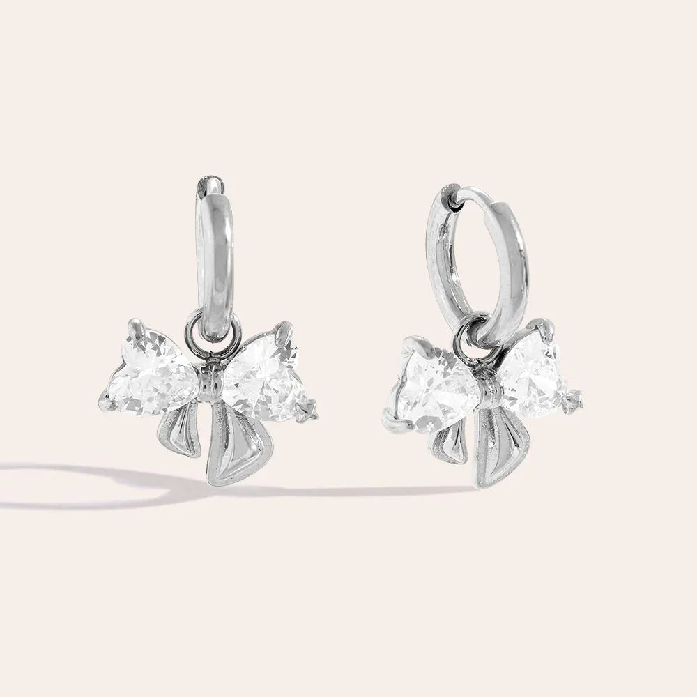 Silberne Creolen-Ohrringe mit glitzernden herzförmigen Anhängern in Schleifenform auf hellem Hintergrund