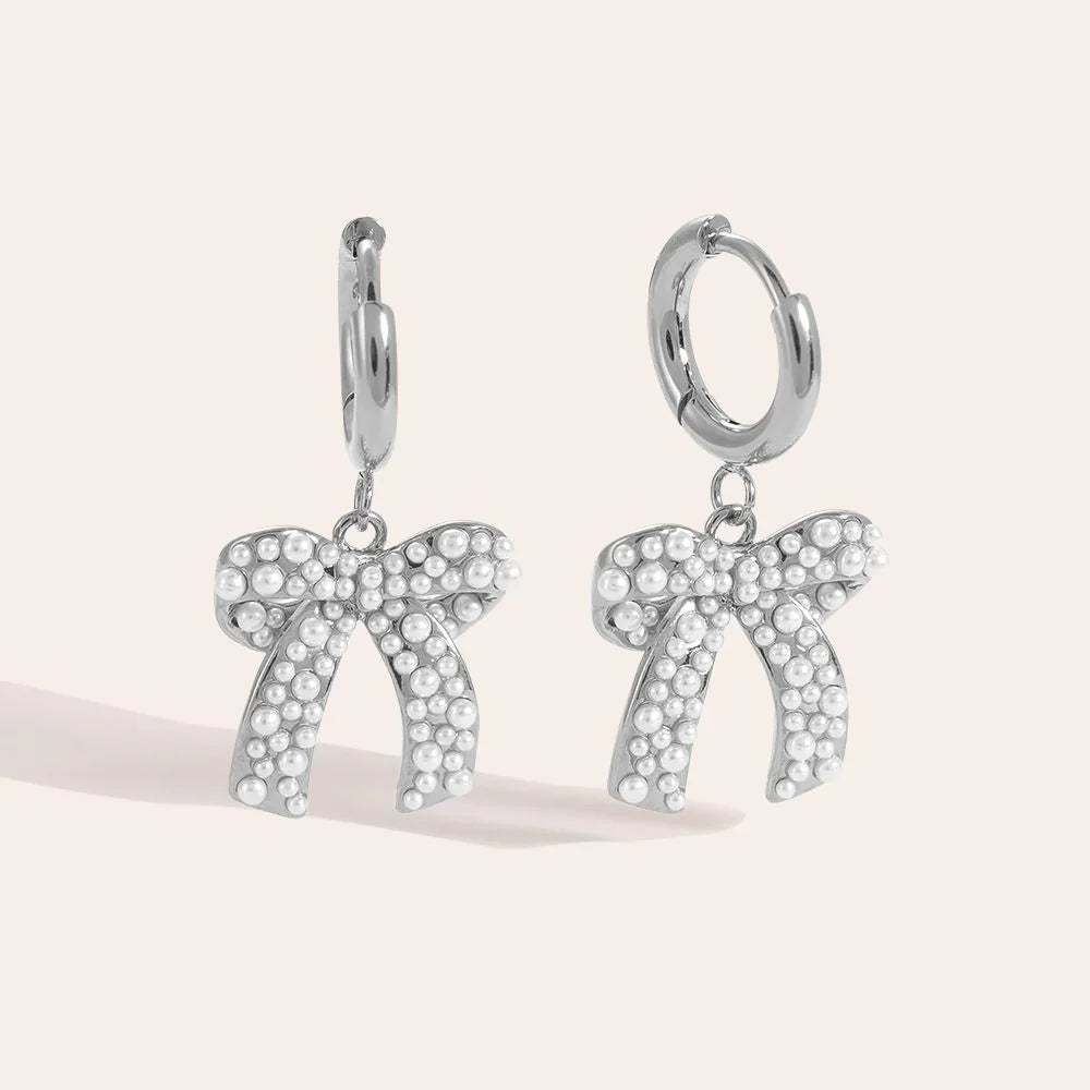 Silberne Creolen-Ohrringe mit weißen Perlen verzierten Schleifen-Anhängern vor hellem Hintergrund
