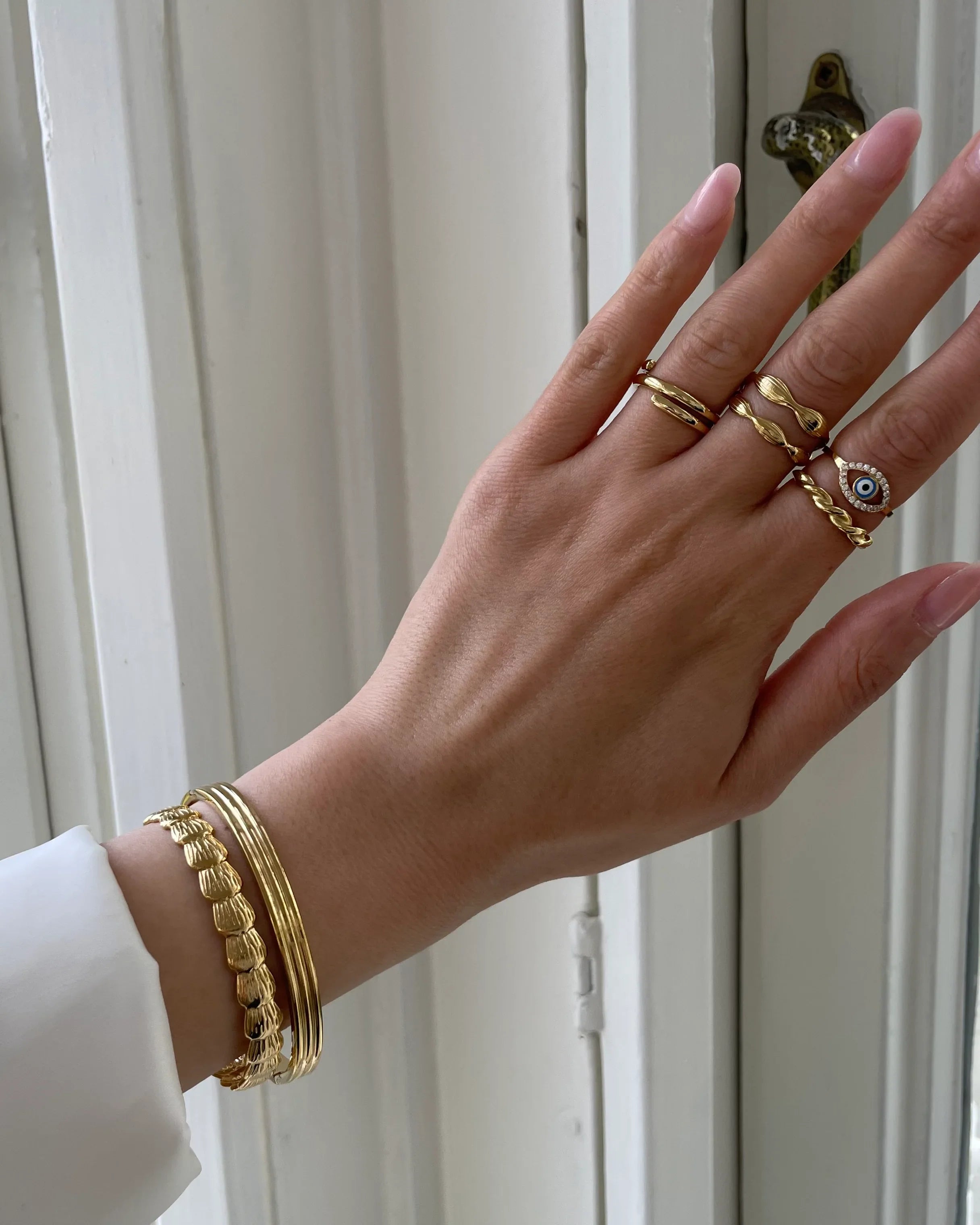 Hand mit goldenen Ringen und Armbändern vor weißer Tür, eleganter Schmuck im Fokus