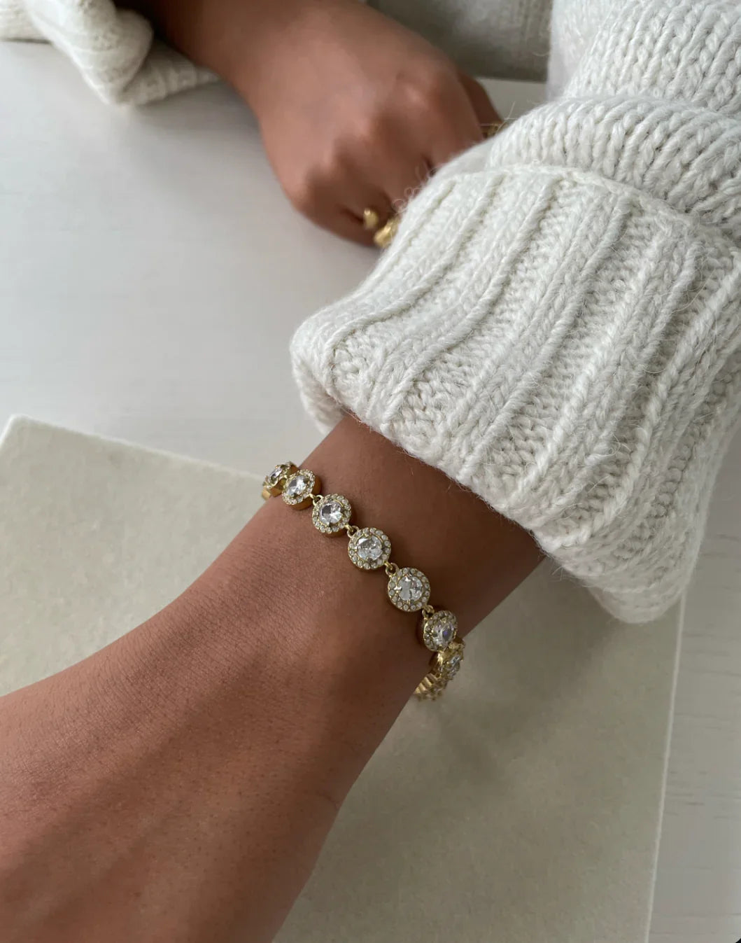 Armband mit goldenen und funkelnden Kristallsteinen am Handgelenk einer Person mit cremefarbenem Strickpullover