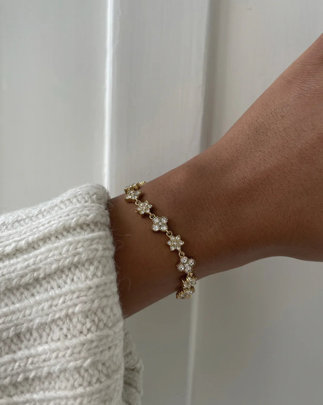 Hand mit goldfarbenem Armband mit funkelnden Blumensteinen vor weißer Wand und weißem Strickpullover