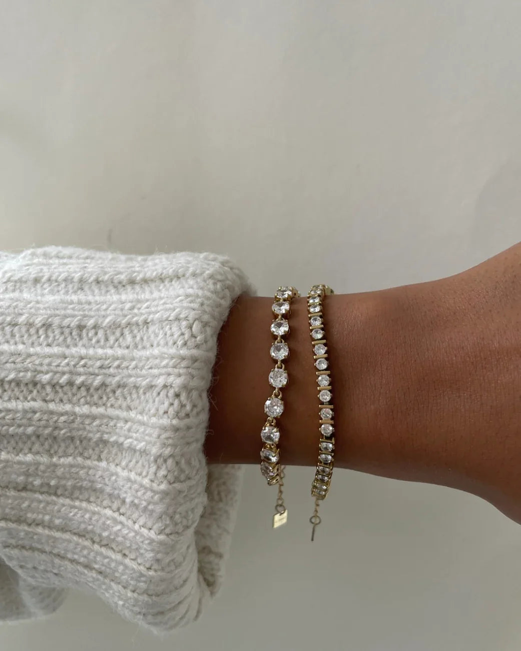 Hand mit zwei goldenen Armbändern mit Strasssteinen und weißem Strickpullover