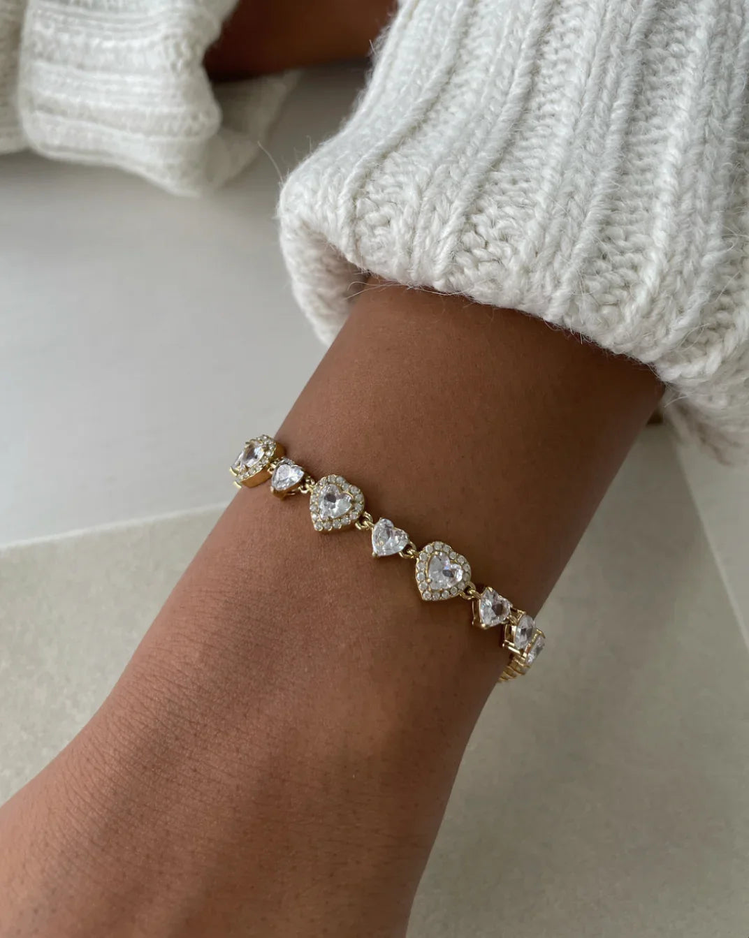 Armschmuck mit herzförmigen und runden Kristallen in Goldfassung am Handgelenk, weiße Strickjacke