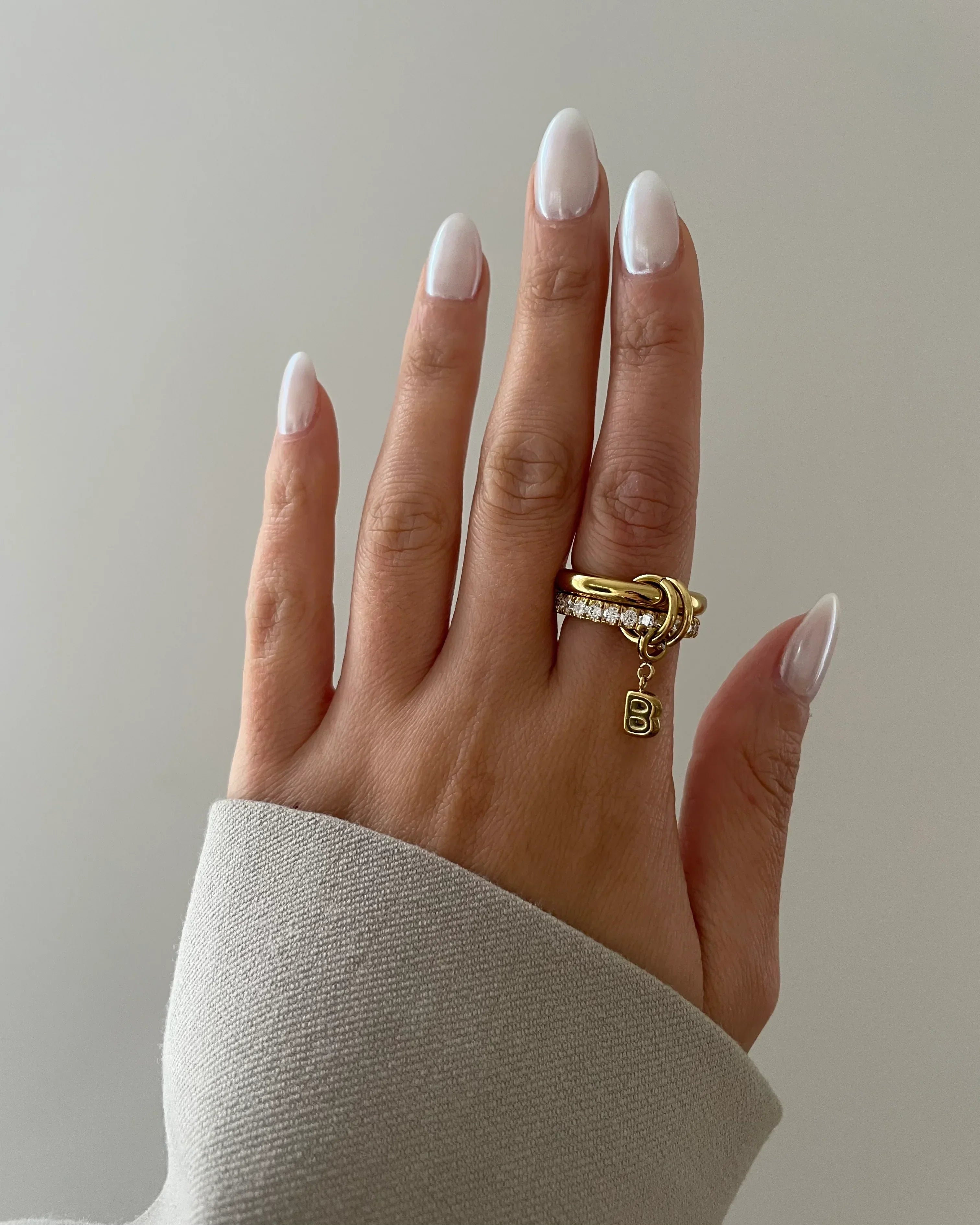 Hand mit hellen, mandelförmigen Nägeln und zwei goldenen Ringen, darunter ein Ring mit Buchstabenanhänger B