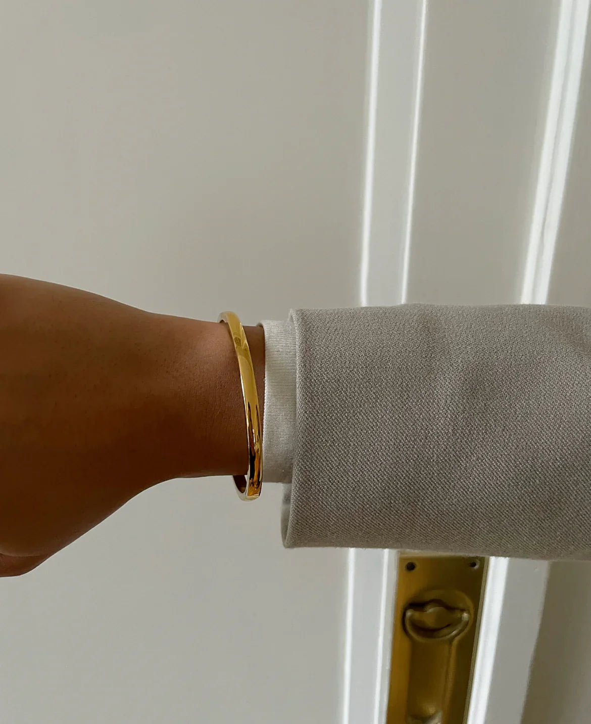 Armgelenk mit schlichter goldener Armspange und grauem Mantelärmel vor weißer Tür