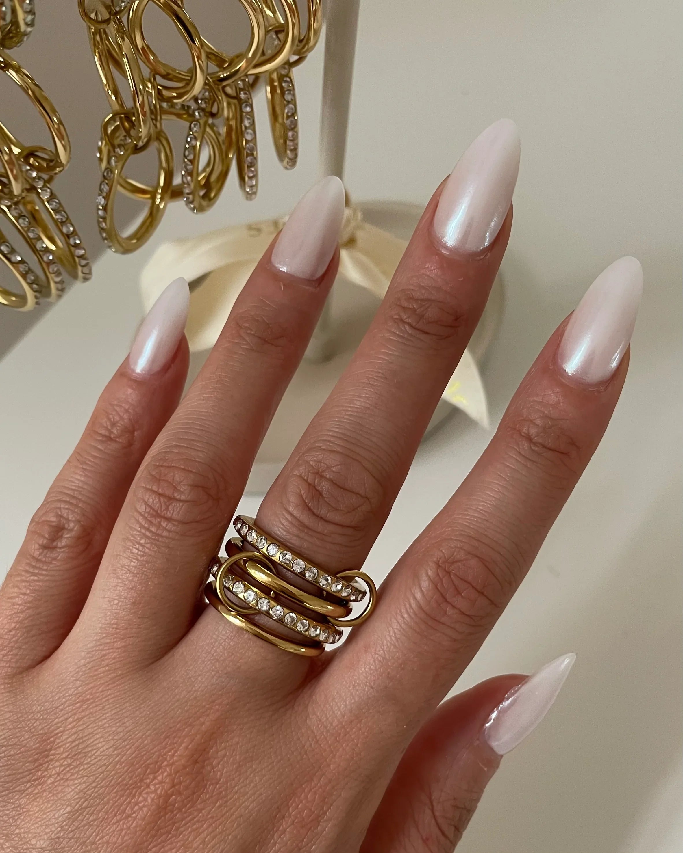 Hand mit langen weißen Nägeln trägt goldenen Ring mit klaren Kristallen, daneben Goldringe mit Kristallen