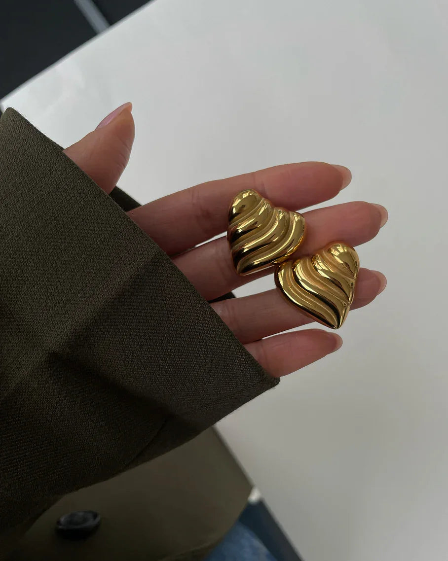 Hand hält zwei goldene, wellenförmige Herz-Ohrringe vor weißem Hintergrund