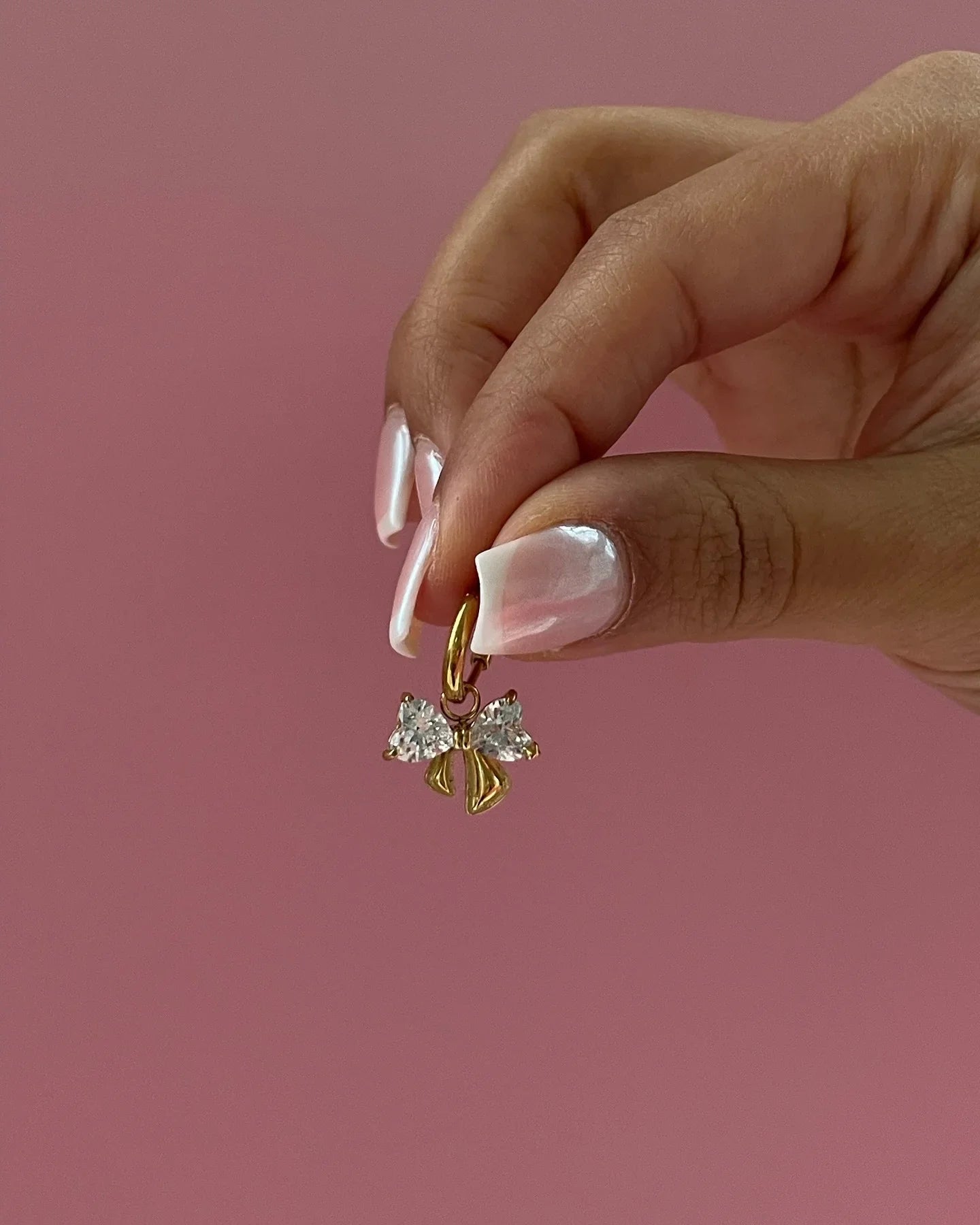 Hand mit gepflegten Nägeln hält goldenen Schleifenanhänger mit Glitzersteinen vor rosa Hintergrund
