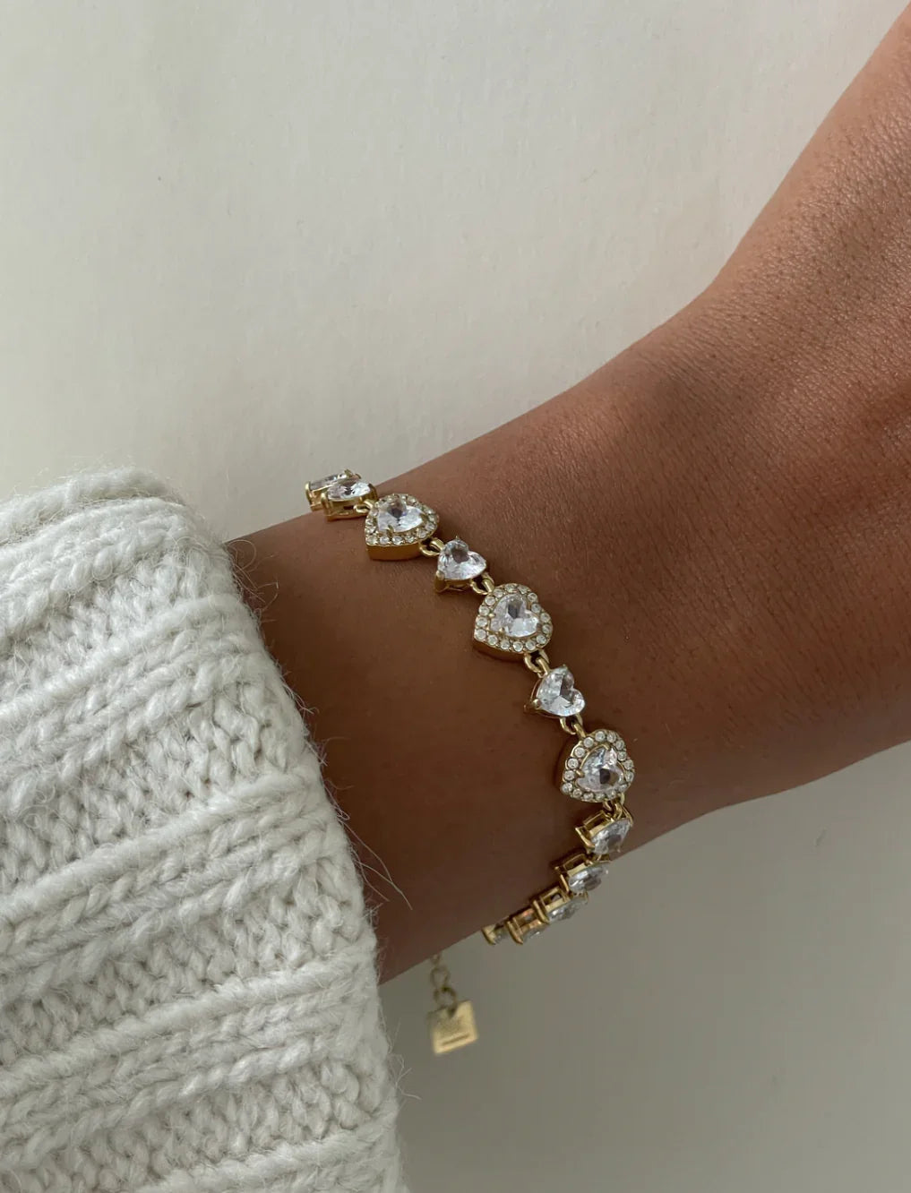 Armband mit goldfarbenen Herzanhängern und weißen Kristallen am Handgelenk vor hellem Hintergrund