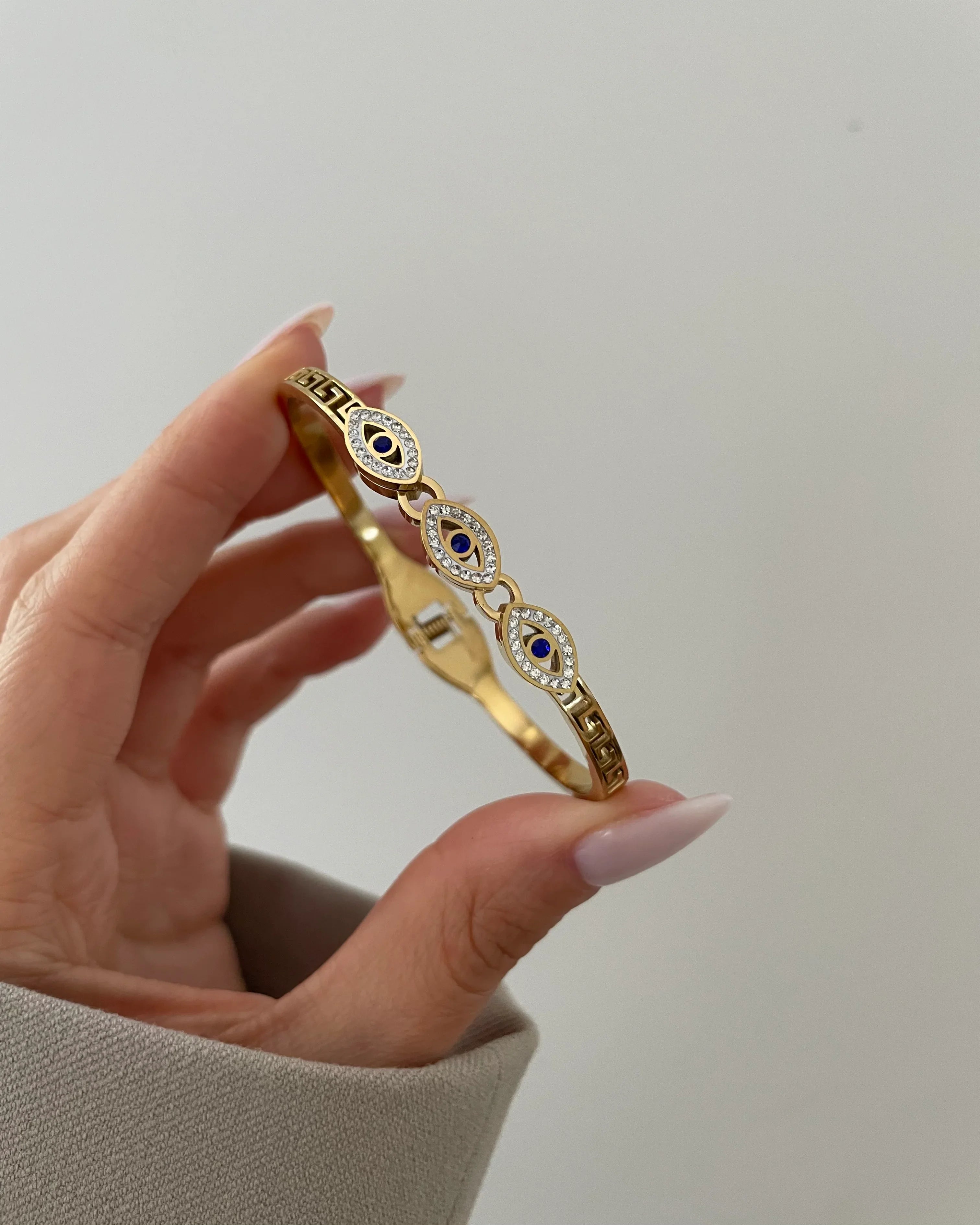 Hand hält goldfarbenes Armband mit drei blauen Augen und Zirkonia-Steinen vor hellem Hintergrund