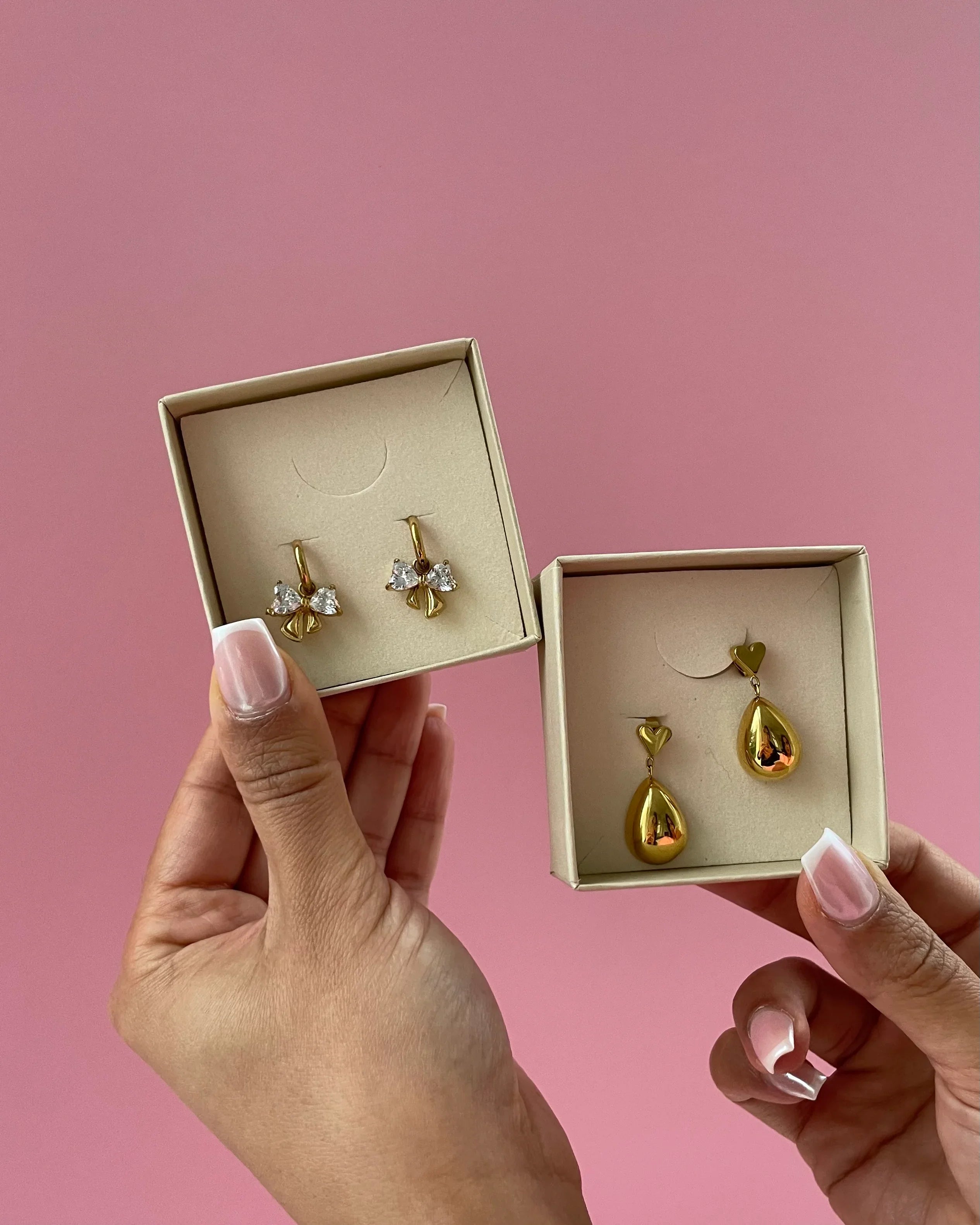 zwei Paar goldene Ohrringe in Geschenkboxen, eine mit Herz- und Tropfenform, die andere mit Kristallschleifen, vor rosa Hintergrund