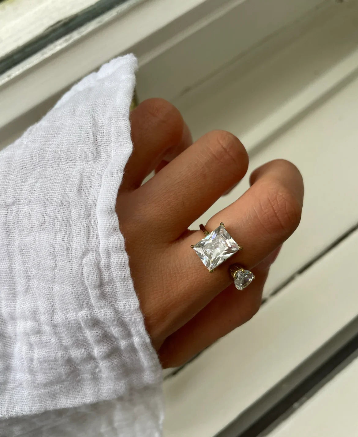 Hand mit weißem Leinenhemd trägt goldenen Ring mit großem quadratischen und kleinem herzförmigen Diamant