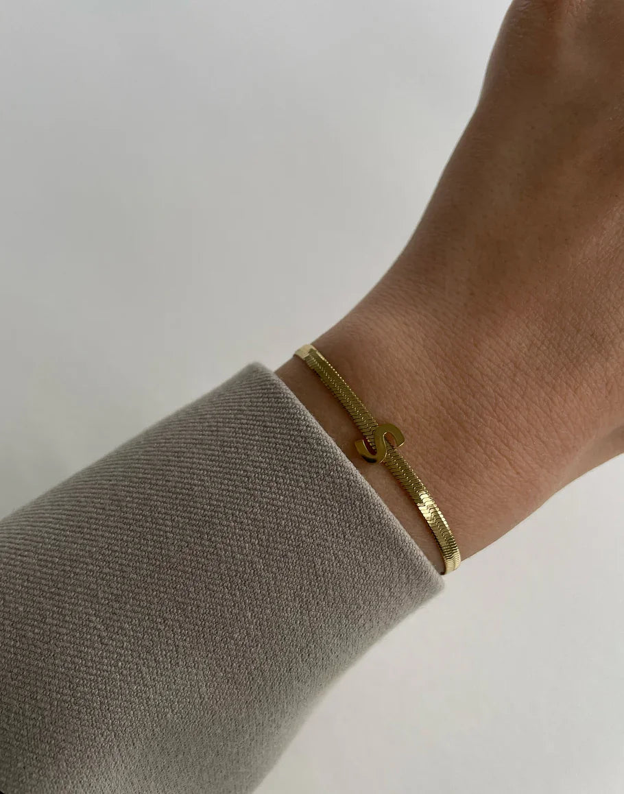 Feines goldenes Armband mit S-Buchstaben-Anhänger am Handgelenk, getragen mit grauem Ärmel