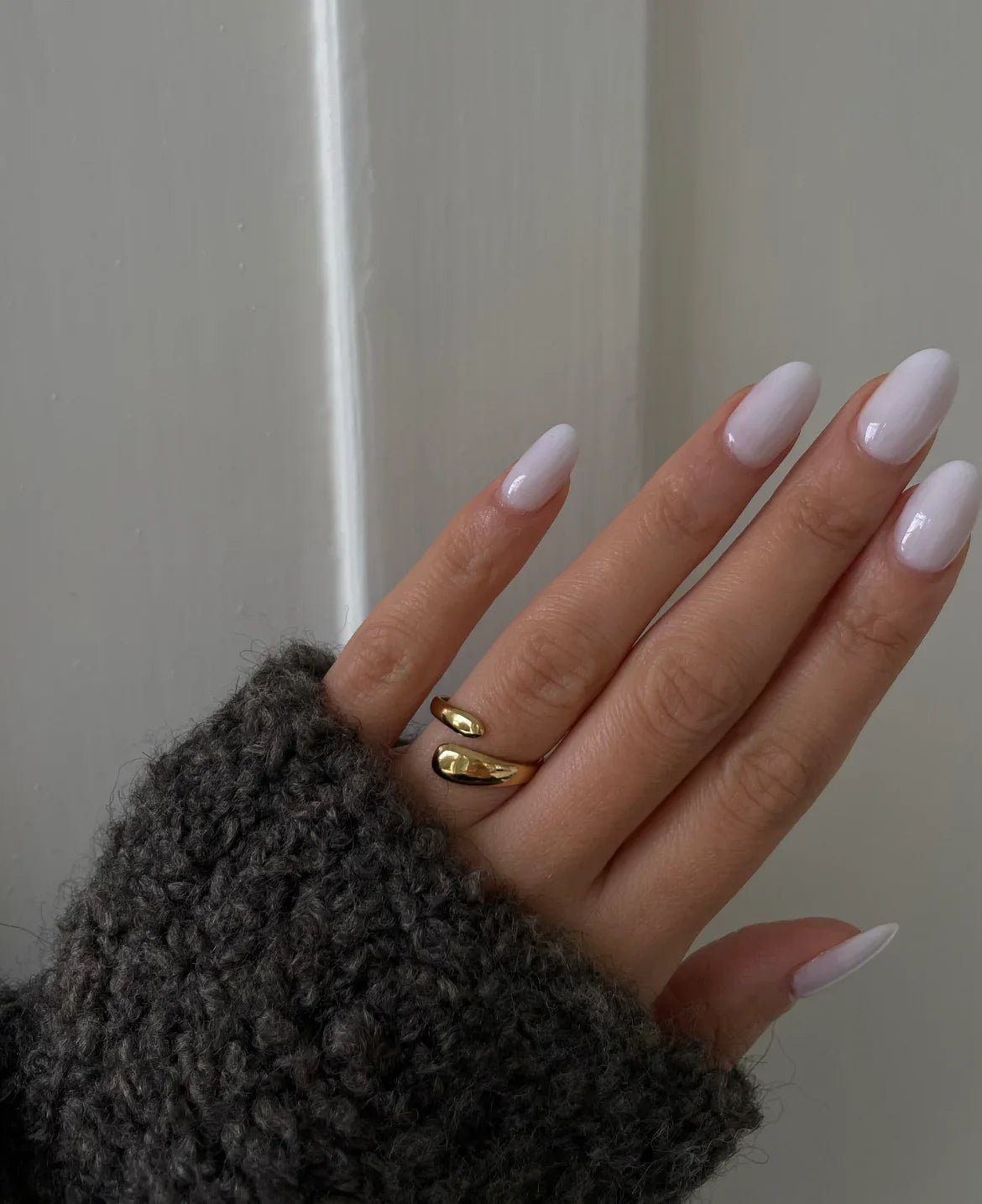 Hand mit weißen, glänzenden Nägeln und goldfarbenem Ring, trägt grauen Strickpullover