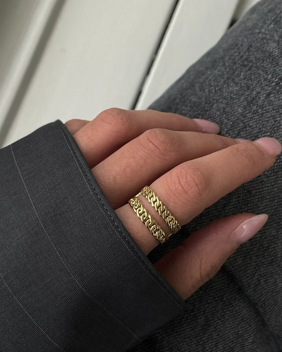 Hand mit zwei goldfarbenen Kettenringen, dunkles Hemd und Jeans im Hintergrund