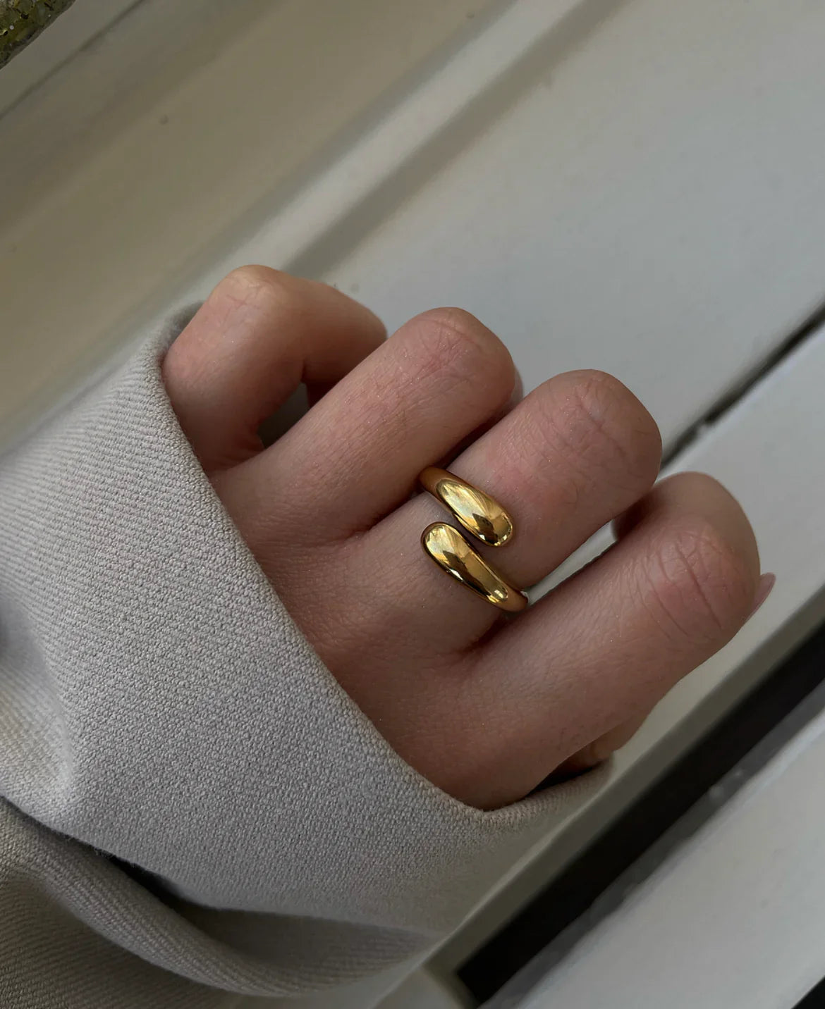 Hand mit goldfarbenem, offenem Ring und grauem Stoffärmel vor hellem Hintergrund
