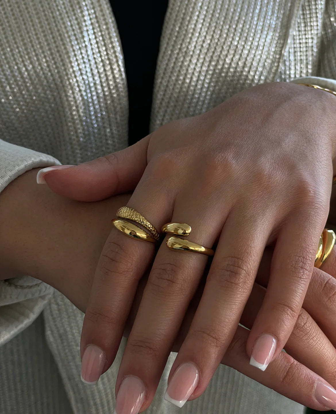 Nahaufnahme von Frauenhänden mit goldenen Ringen und French Maniküre vor beigem Strickpullover