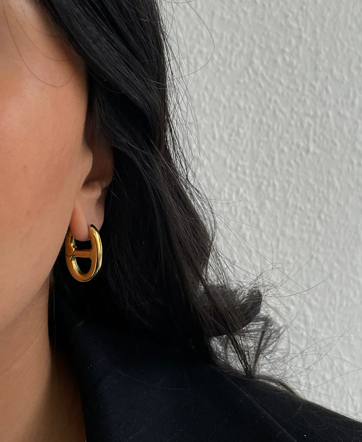 Nahaufnahme goldenes Hufeisen-Ohrring an Frau mit dunklem Haar vor weißer Wand