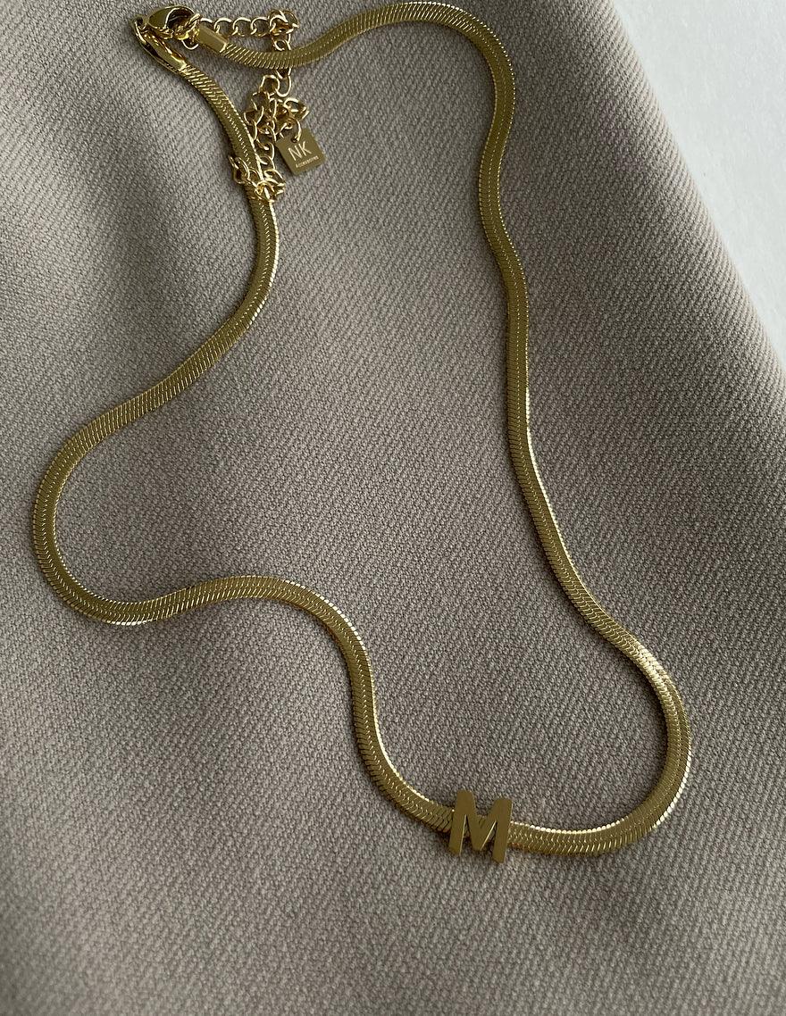 Goldfarbene Halskette mit Buchstabe M auf beigem Stoffhintergrund