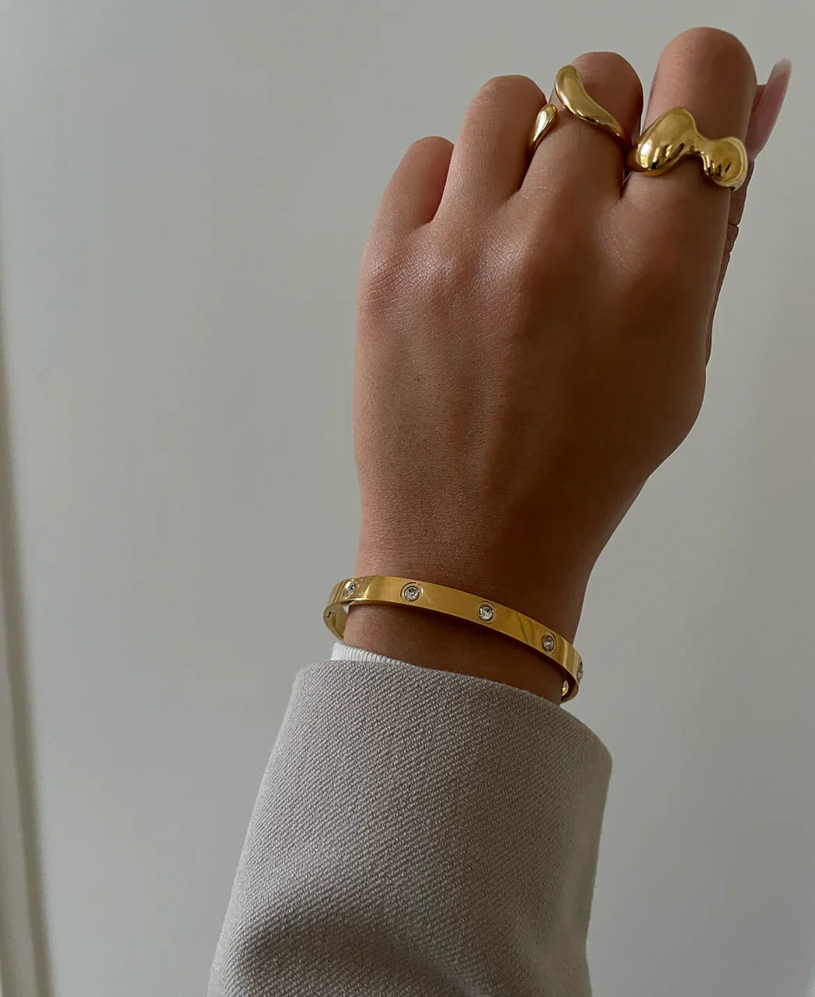 Hand mit goldfarbenem Armreif mit Kristallen und zwei goldenen Ringen vor grauem Hintergrund