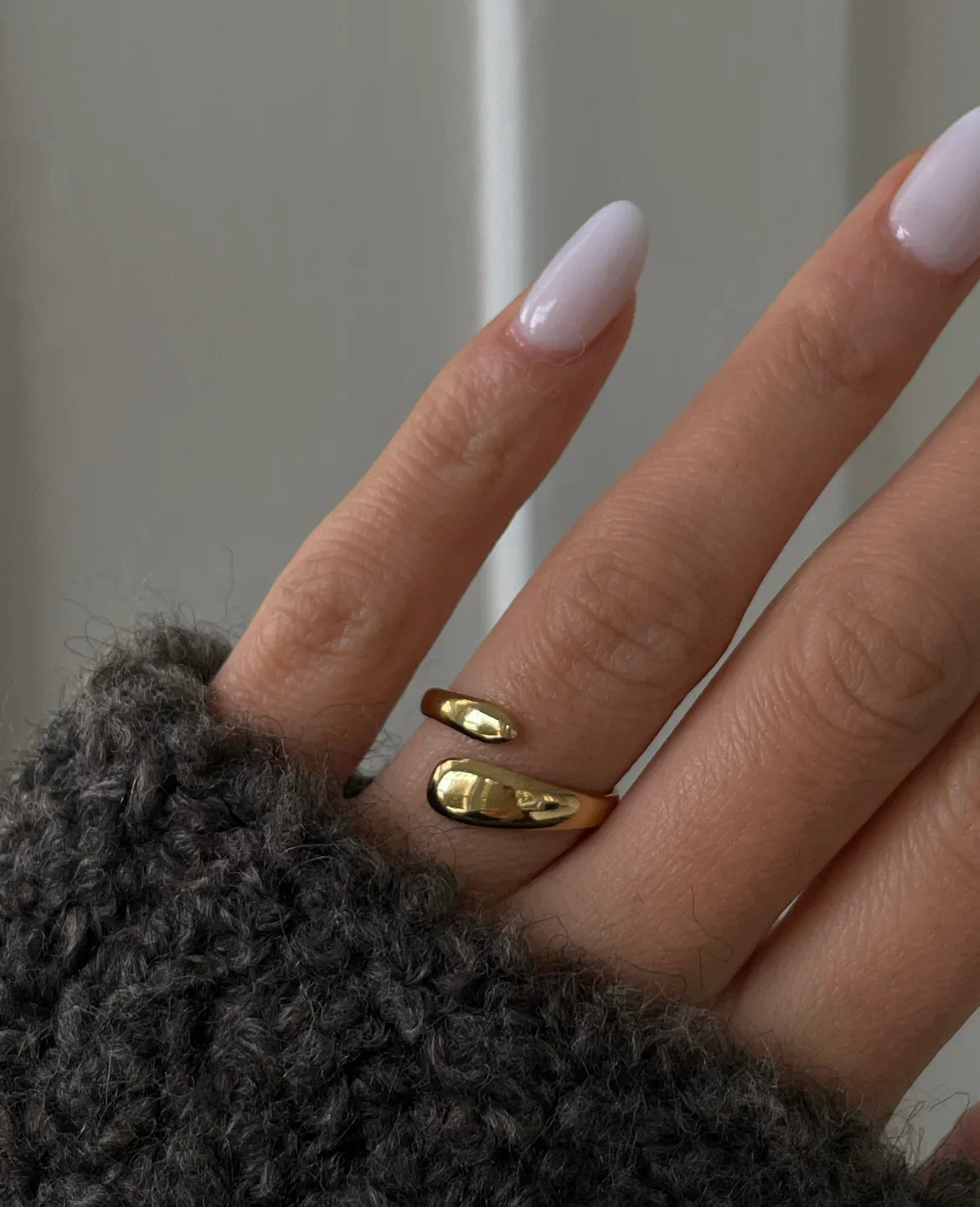 Hand mit elegantem offenem goldenen Ring und pastellfarbenem Nagellack, trägt grauen Pullover