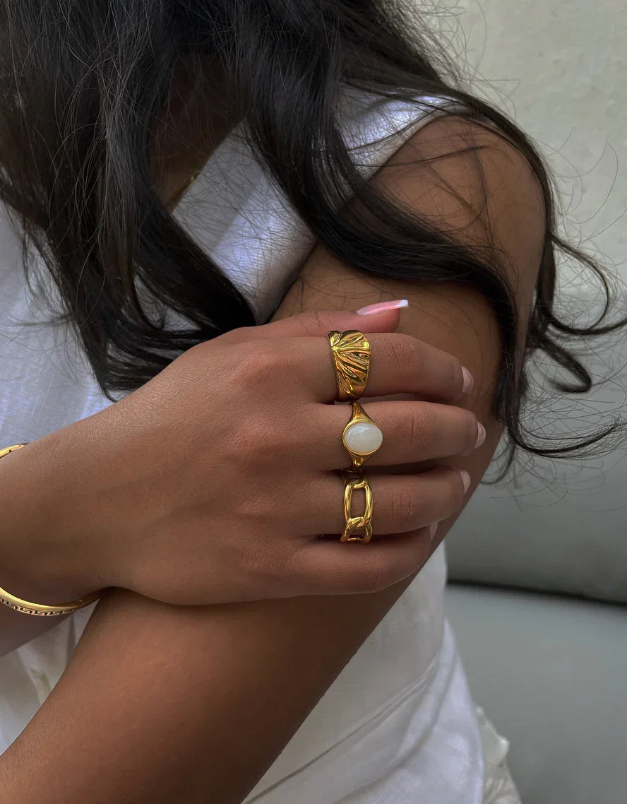 Nahaufnahme eines Arms mit drei goldenen Ringen und Armband, helle Haut, dunkle Haare, weißes Kleid