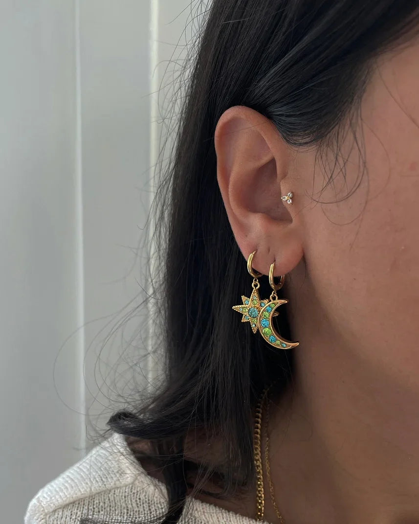 Nahaufnahme eines Ohrs mit goldenen Creolenohrringen in Stern- und Mondform mit grünen und blauen Edelsteinen