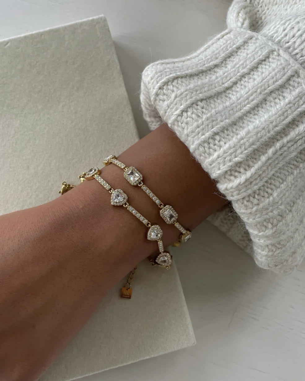 Armgelenk mit zwei goldenen Armbändern mit glitzernden Herz- und Rechteck-Diamanten an hellem Strickpullover