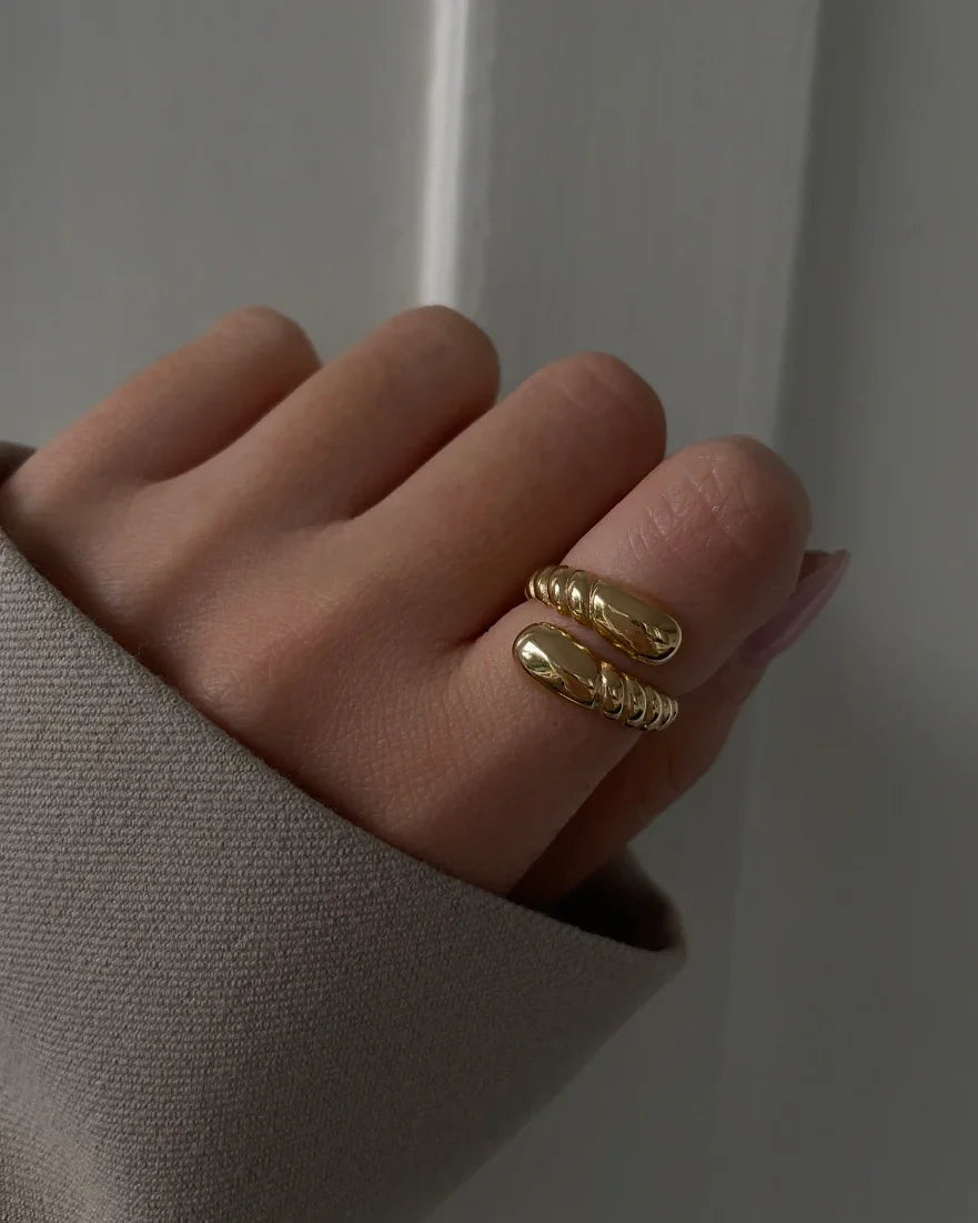 Hand mit goldfarbenem, gewickeltem Offener Ring und beigem Stoffärmel auf grauem Hintergrund