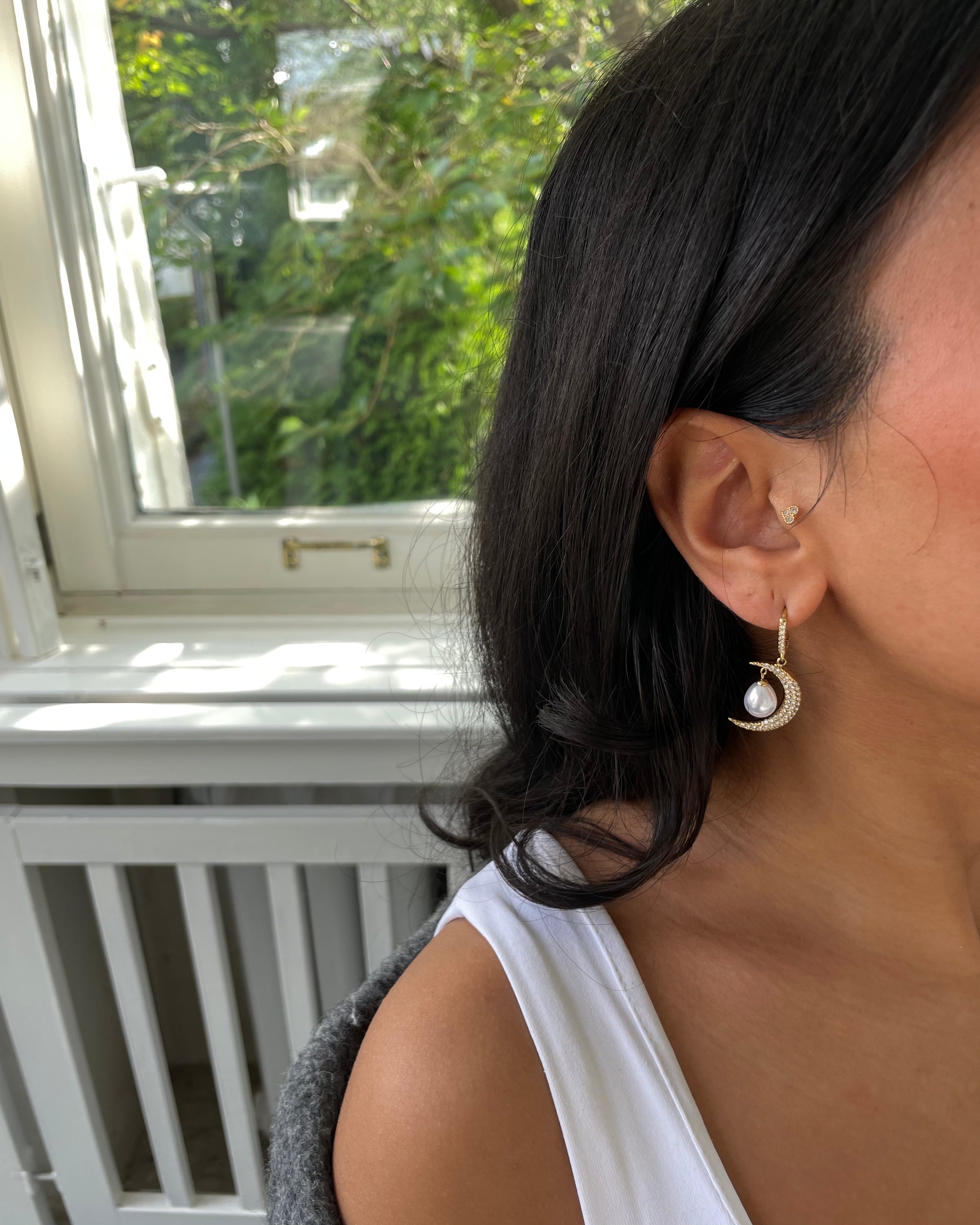 Frau mit schwarzem Haar trägt goldene Mond-Ohrringe mit Perle im hellen Raum vor Fenster