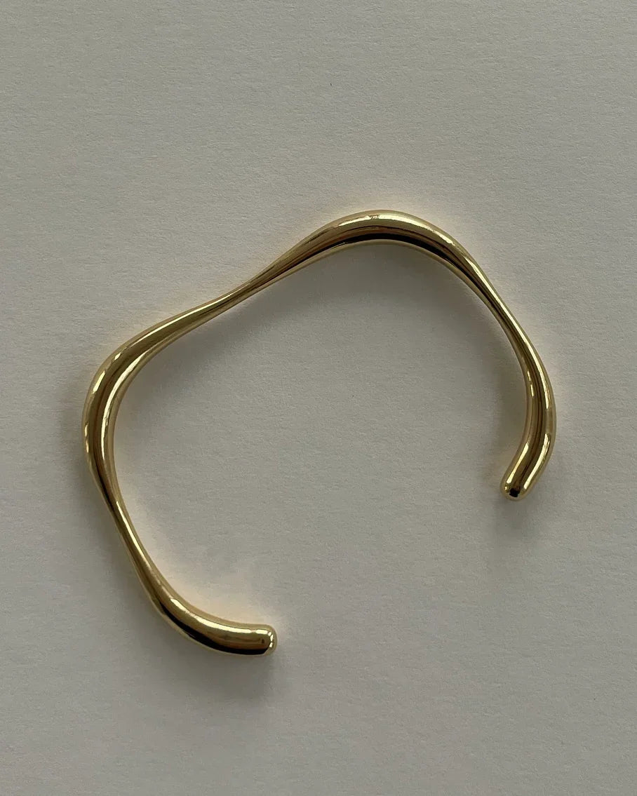 Minimalist goldfarbener offener Armreif mit unregelmäßiger, organischer Form auf hellem Hintergrund