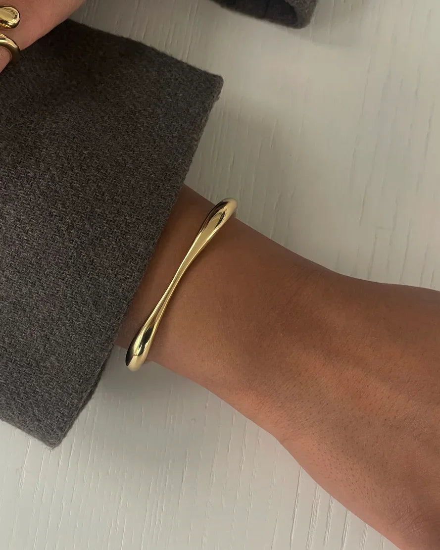 Handgelenk mit modernem, glänzendem goldfarbenem Armreif und grauem Wollpullover auf hellem Holztisch