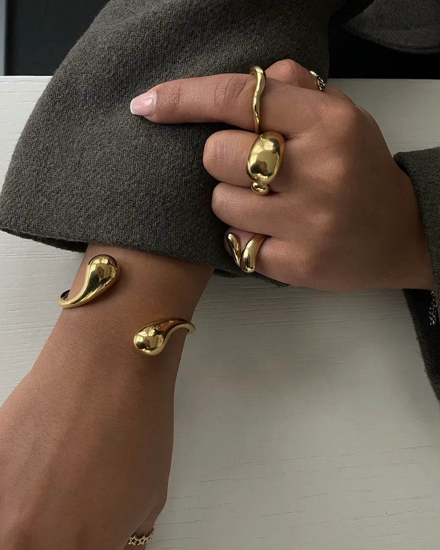 Hand mit goldenen organisch geformten Ringen und Armreif auf weißem Untergrund, brauner Pullover