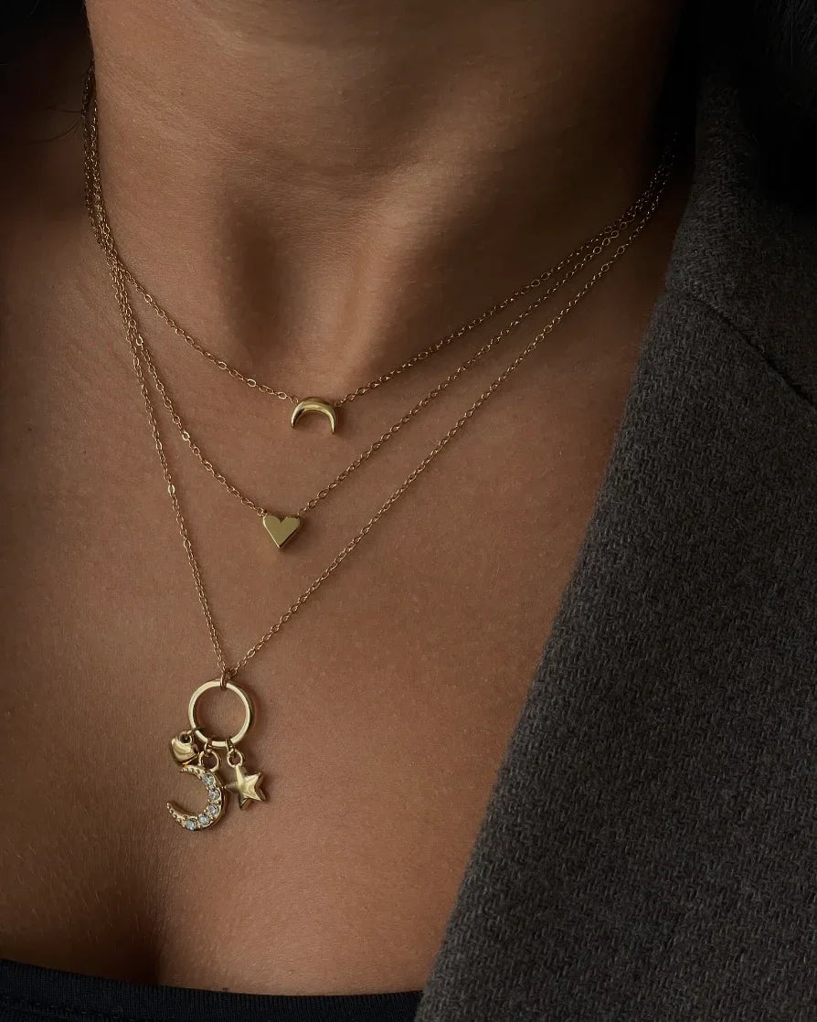Dreifache goldene Halsketten mit Mond-, Herz- und Sternanhängern auf heller Haut und grauem Mantel