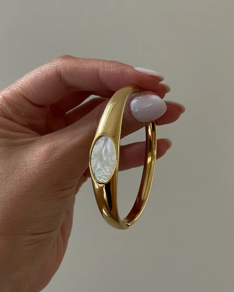 Hand hält goldfarbenen Armreif mit weißem Perlmutt-Einsatz vor beigem Hintergrund