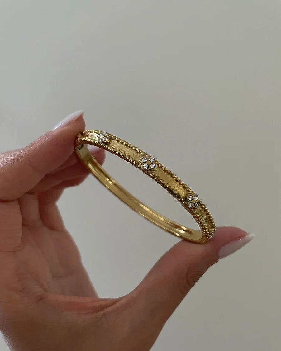 Hand hält goldfarbenen Armreif mit kleinen Kristallblumen auf hellgrauem Hintergrund