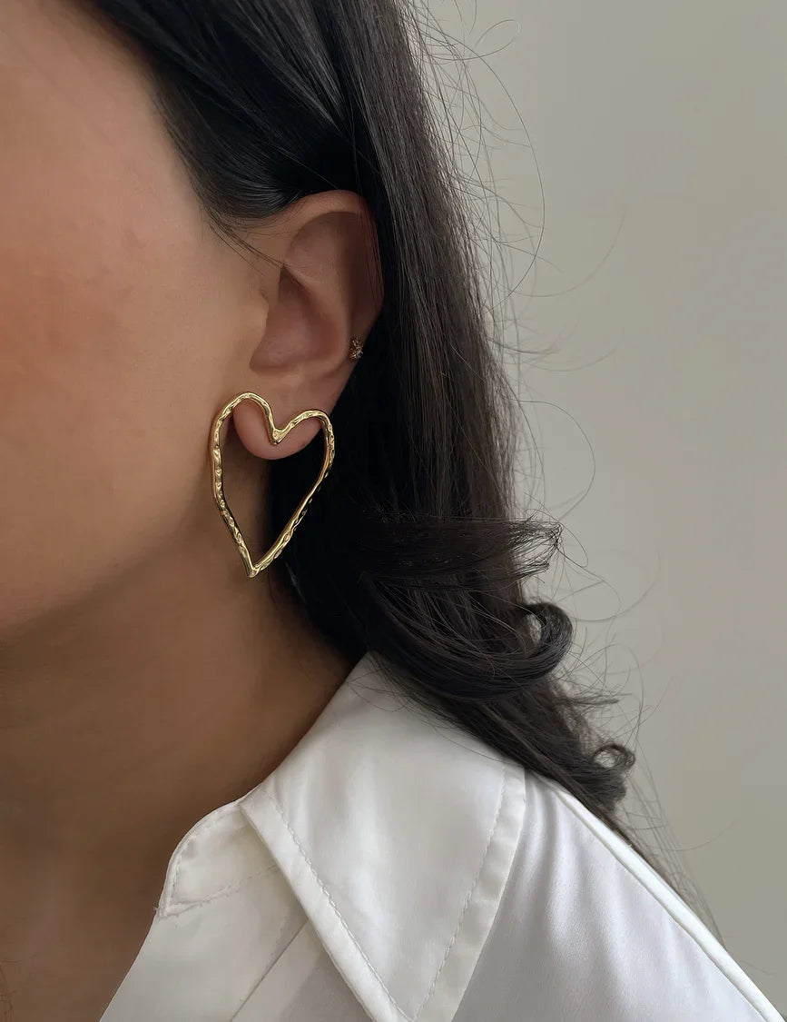 Frau mit herzförmigem Goldohrring und weißer Bluse vor neutralem Hintergrund