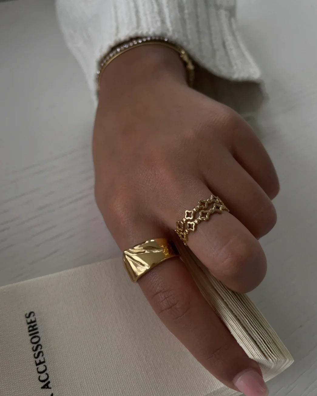 Hand mit goldenen Ringen und weißem Pullover, hält Buch mit Titel ACCESSOIRES