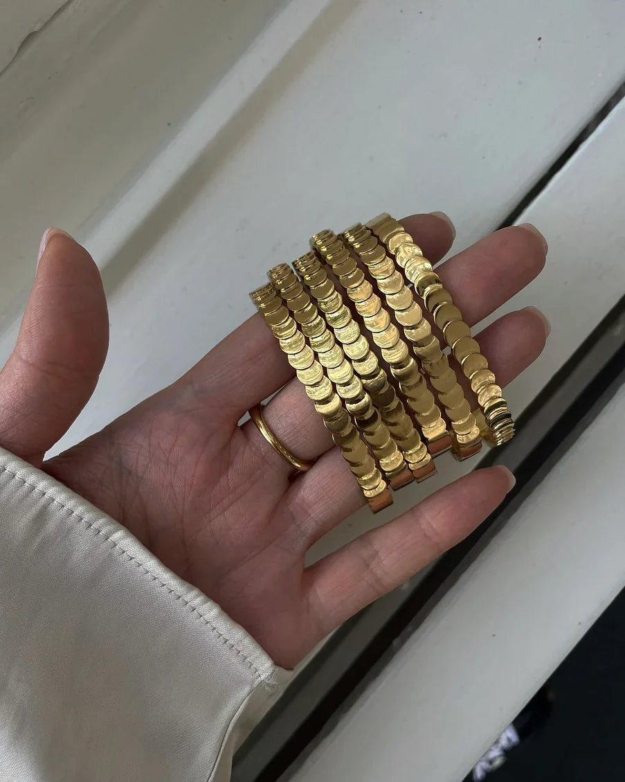 Hand mit goldfarbenem Armband aus runden Plättchen vor hellem Hintergrund, weiße Manschette sichtbar