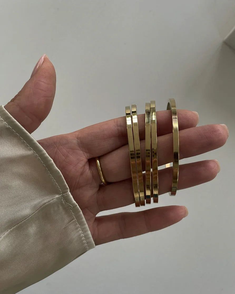 Hand hält fünf schmale goldene Armreifen vor hellem Hintergrund, helle Bluse sichtbare Manschette