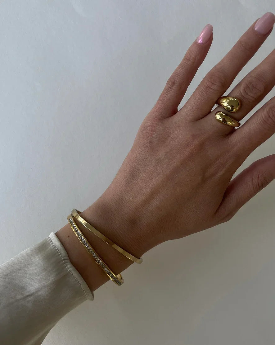 Hand mit goldfarbenem Ring und zwei Armbändern, eines mit Kristallen, vor weißem Hintergrund