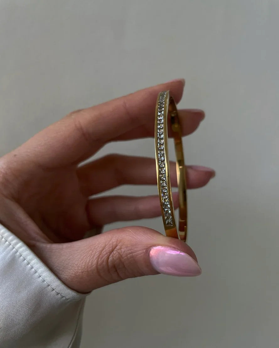 Hand hält goldfarbenes Armband mit funkelnden Kristallen vor neutralem Hintergrund