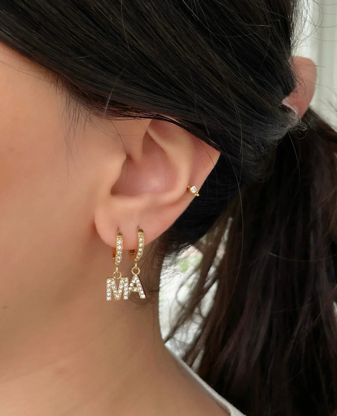 Ohr mit goldenen Creolen und Anhängern mit Buchstaben MA, feines Ohrpiercing mit Kristall