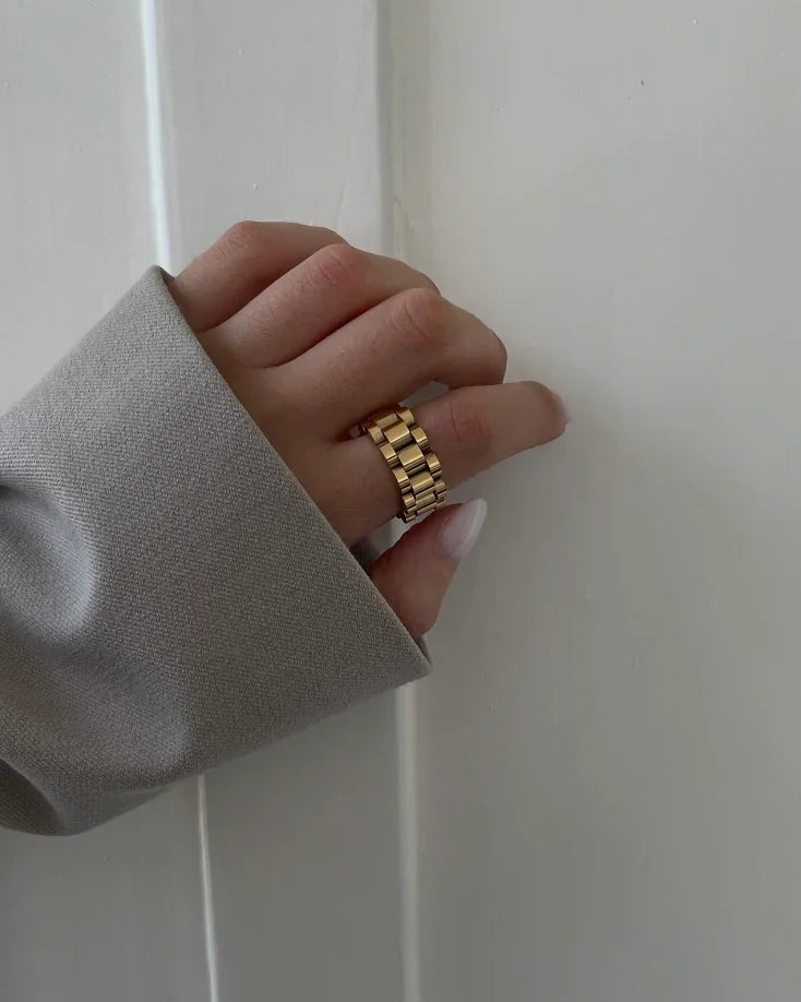 Hand mit goldfarbenem, breitgliedrigem Ring und grauem Ärmel vor weißer Wand