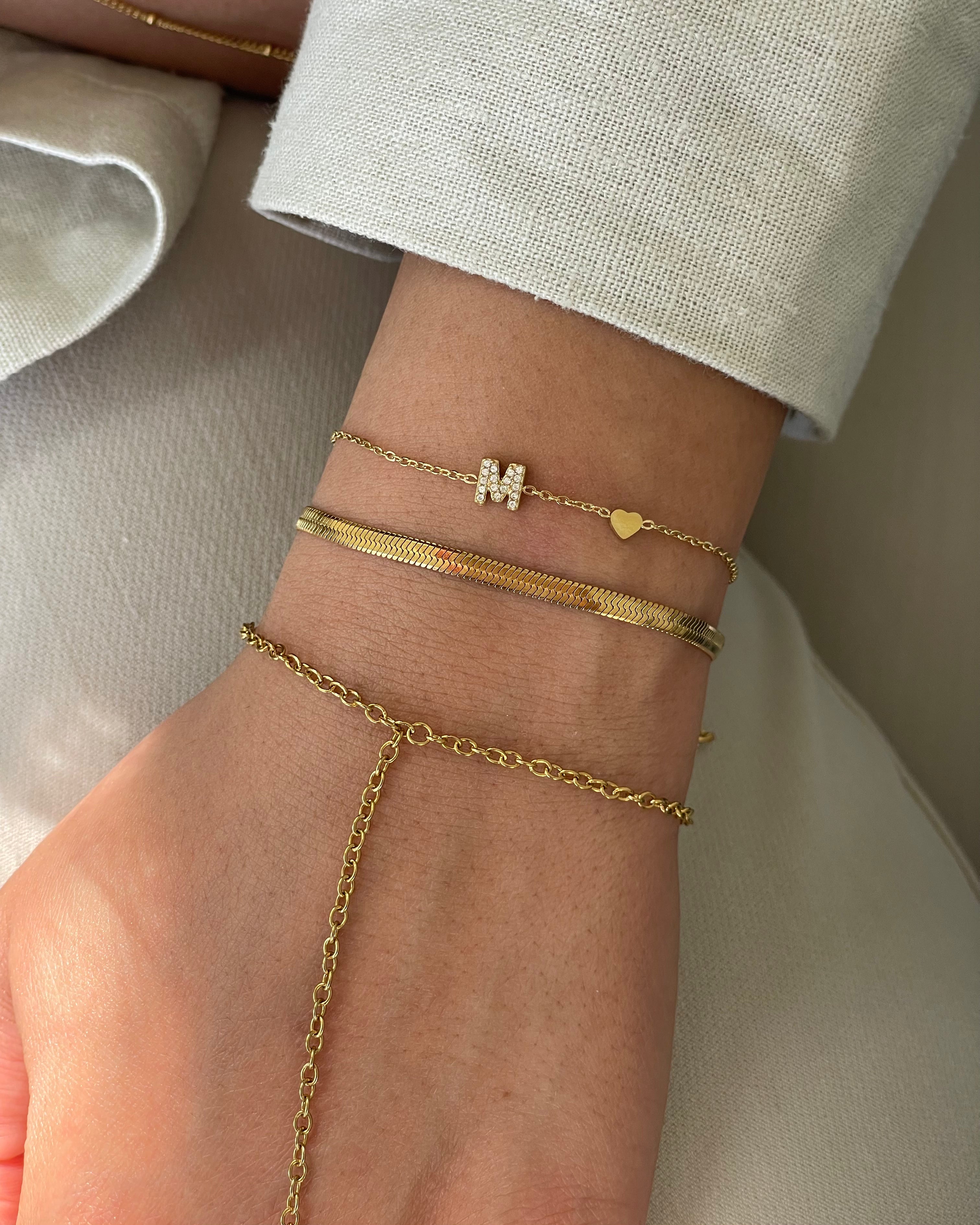 Nahaufnahme eines Handgelenks mit drei goldenen Armbändern, darunter eins mit Buchstabe M und Herz