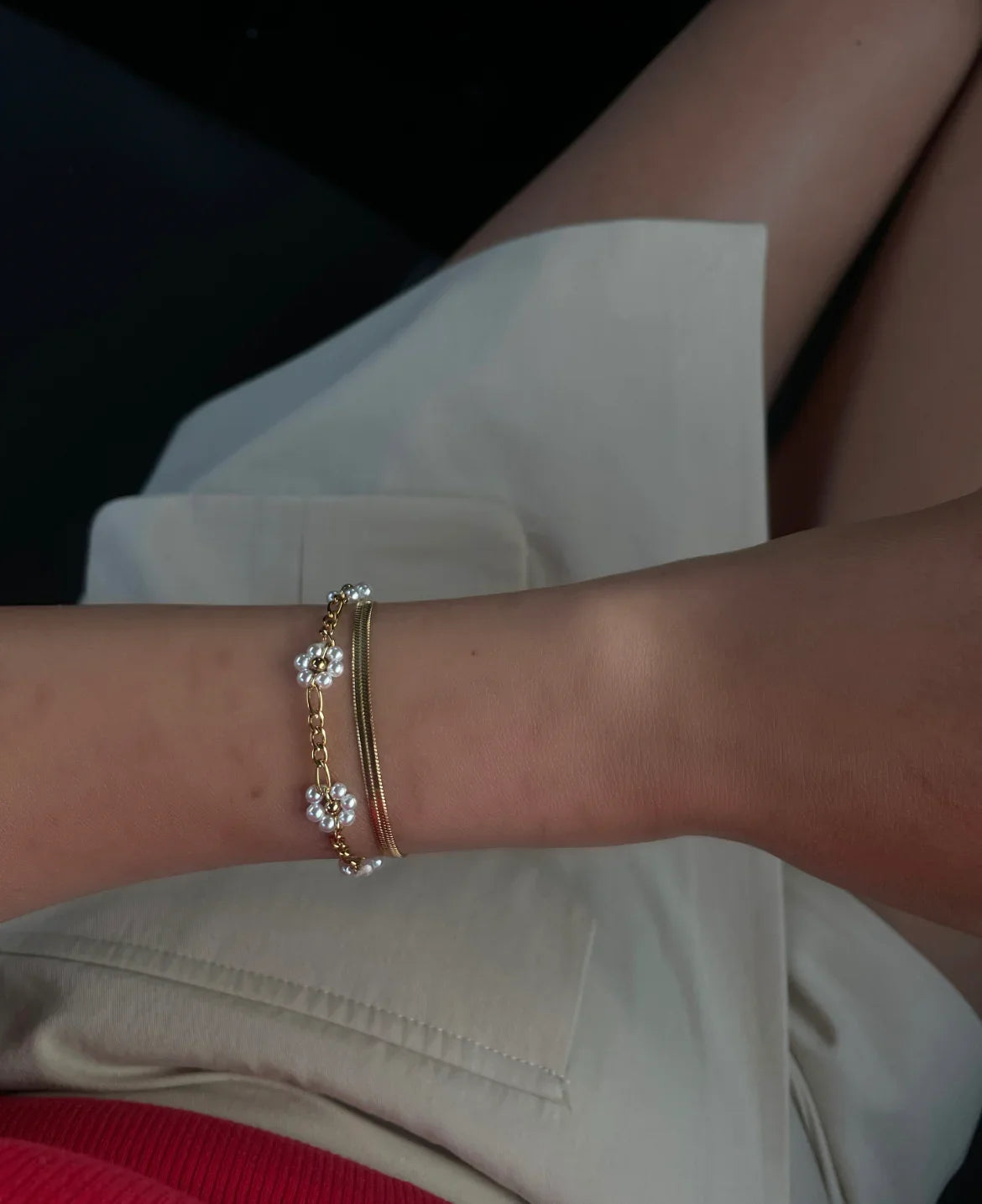 Armband mit goldener Kette und weißen Perlenblumen am Handgelenk einer Person in hellem Rock und rotem Oberteil
