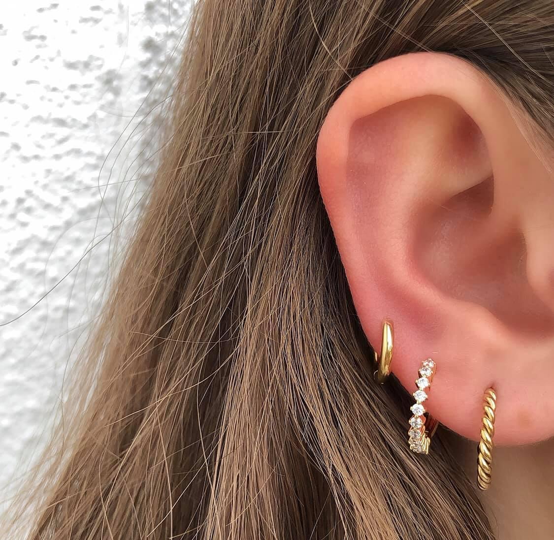 Nahaufnahme eines Ohrs mit drei verschiedenen goldenen Ohrringen an einem braunhaarigen Modell