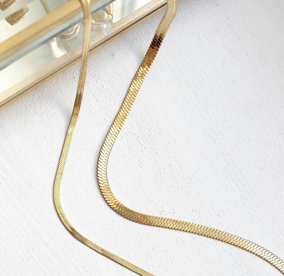 Goldfarbene flache Schlangenkette auf weißem Untergrund neben goldenem Spiegelrahmen