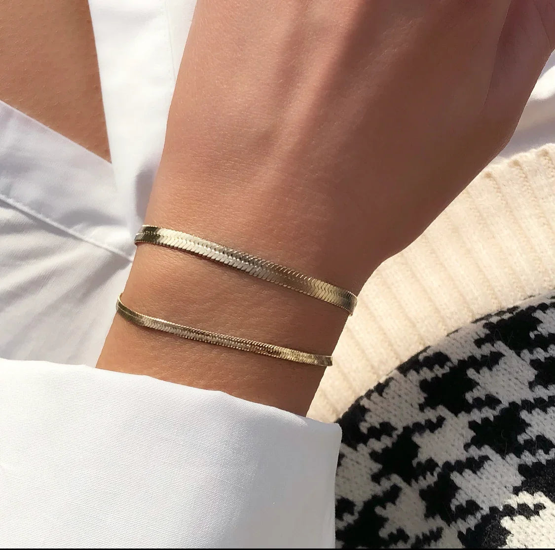 Hand mit zwei goldenen, feinen Armbändern auf weißem Hemd und schwarz-weißem Muster