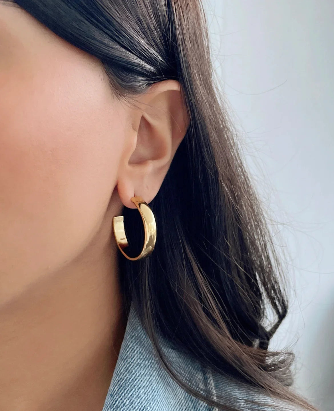 Frau trägt glänzende goldene Creolen Ohrringe, Nahaufnahme, dunkelbraune Haare, hellblauer Jeanskragen